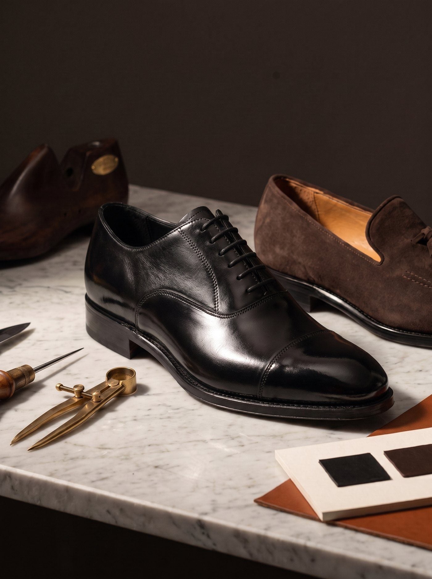 Oxford in abrasivato nero e mocassino in camoscio su un piano di marmo di Carrara, con lesina e compasso in ottone e una cartella di campioni di pelle